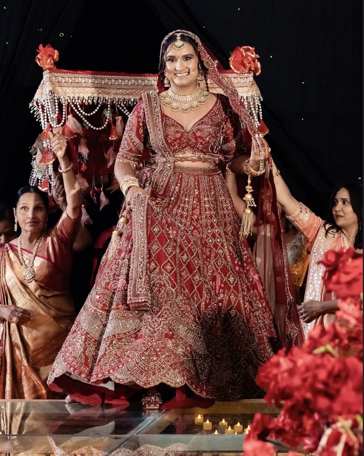 Bride in a red lehenga walking under a floral canopy.